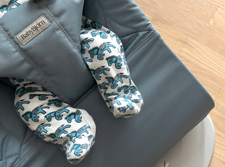 Babybjörn baby shoes with blue dinosaur pattern on a gray chair, indoor setting.